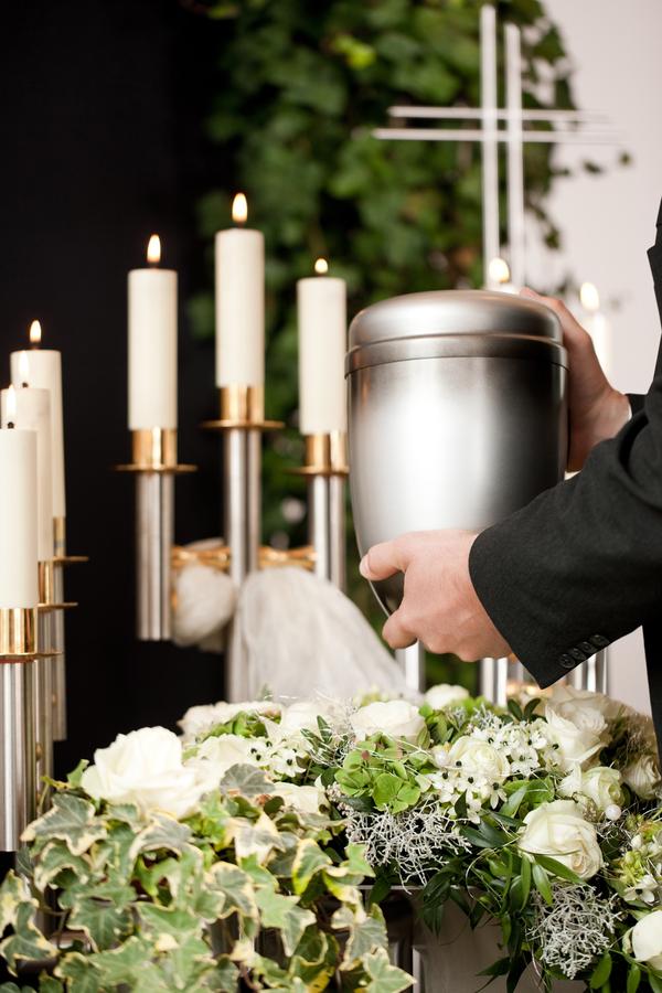Hand, die eine silberne Urne über einem Blumengesteck mit weißen Rosen und Kerzen auf einem Tisch platziert.