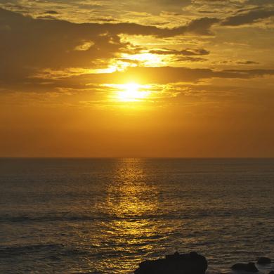 Sonnenuntergang über dem Meer, das sanftes goldenes Licht reflektiert, mit silhouettierten Felsen im Vordergrund.